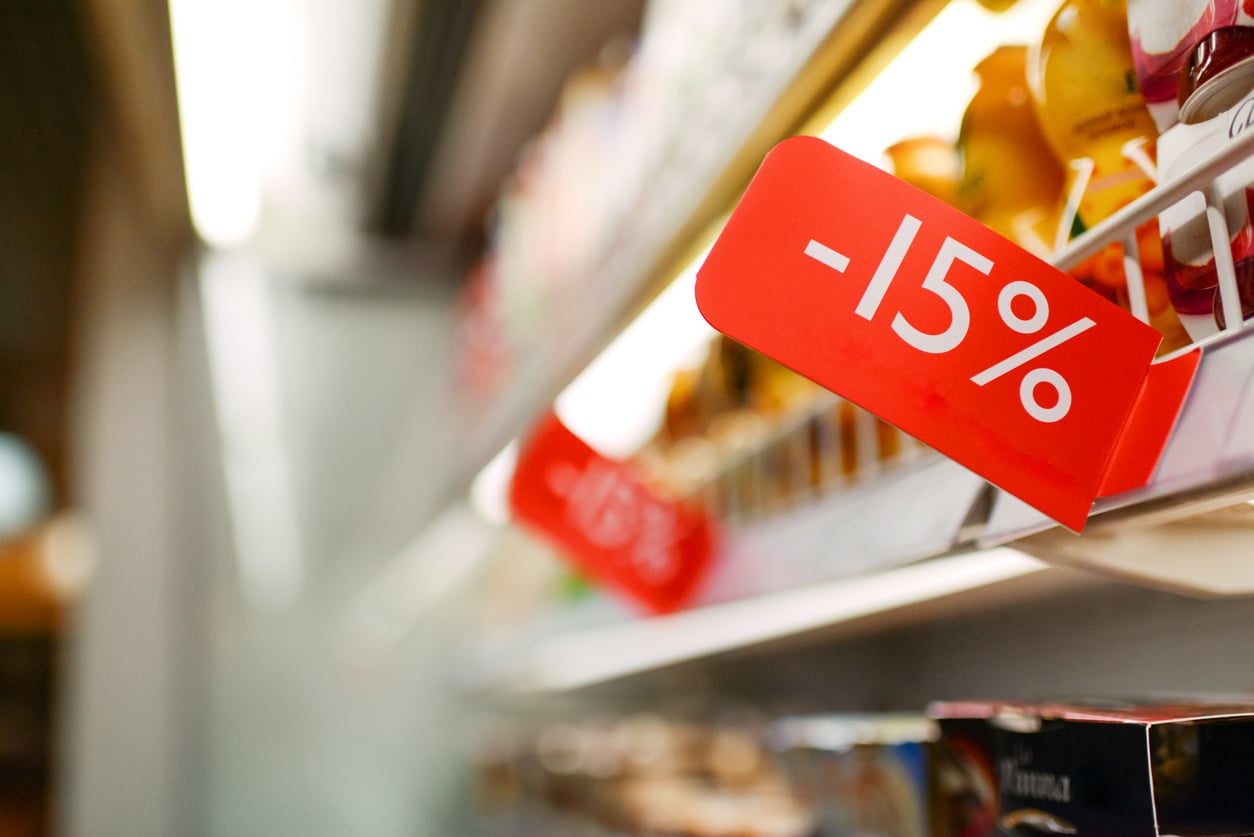 A red sign on a grocery store shelf displays “-15%,” indicating a discount on nearby products and encouraging shoppers to reflect on their grocery habits. Other similar signs are visible further down the aisle.