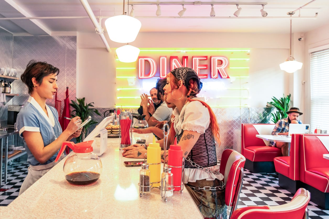 A waitress takes an order from two people at a retro diner counter, their laughter hinting at vacation excitement. A neon “DINER” sign glows in the background as a man relaxes in a booth on the right. The decor features red booths, checkered floor, and plants.