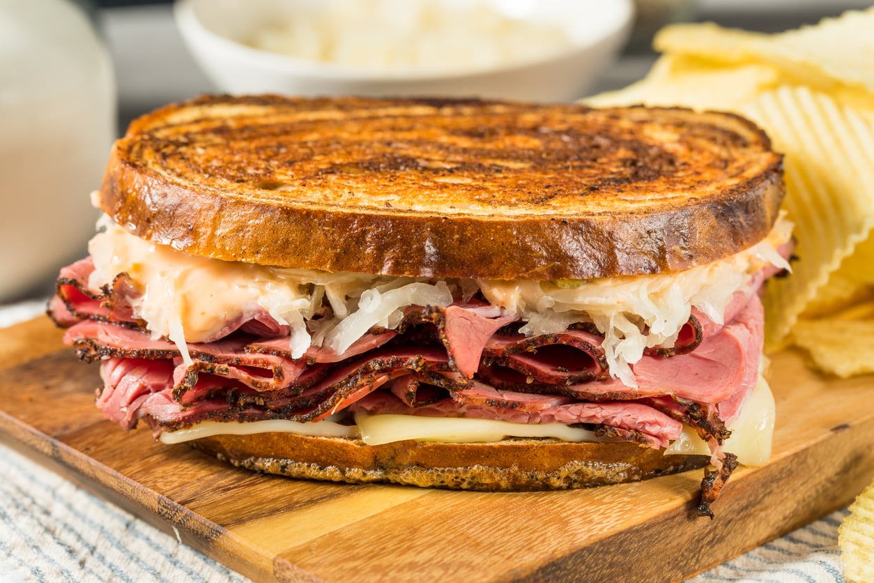 A Reuben sandwich with layers of pastrami, sauerkraut, melted Swiss cheese, and dressing on toasted rye bread, served on a wooden board with potato chips in the background.