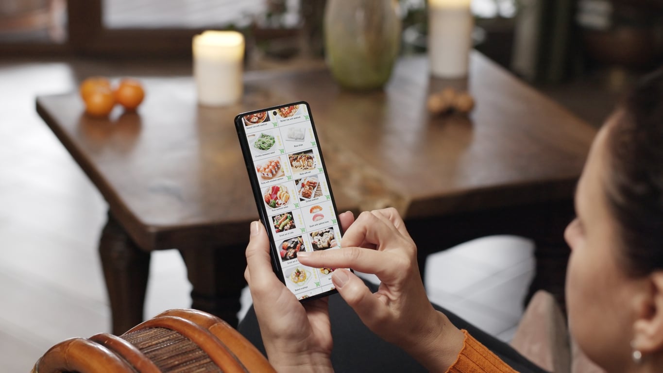 A person browses a food delivery app on a smartphone while sitting indoors, with a wooden table, candles, and fruit visible in the background.