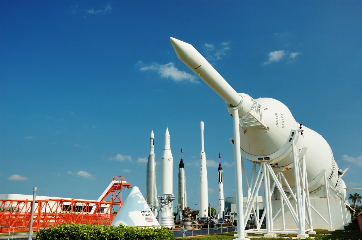 An outdoor display of historic rockets stands under a clear blue sky at a space center, with a large white rocket in the foreground and several smaller rockets and structures in the background.