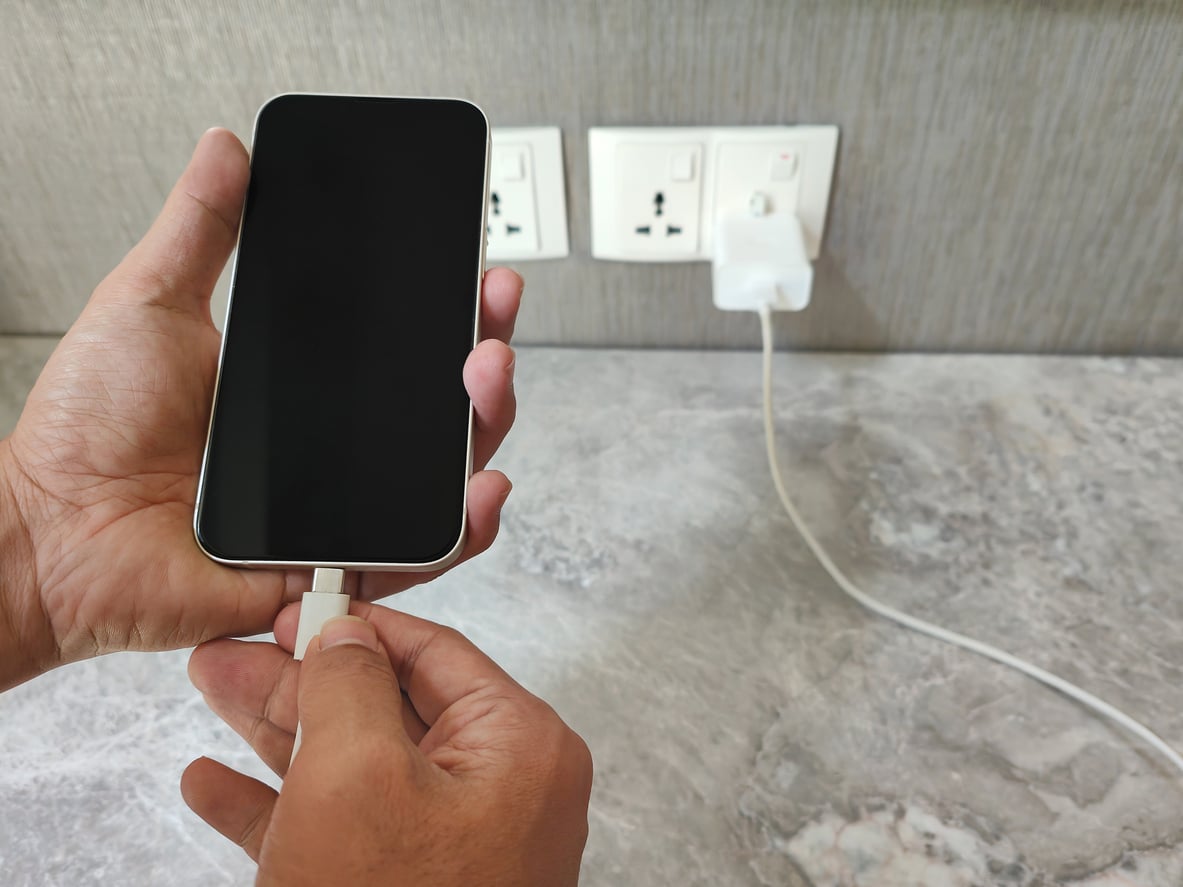 A person is plugging a charging cable into a smartphone, with the other end connected to a wall socket on a gray marble countertop.