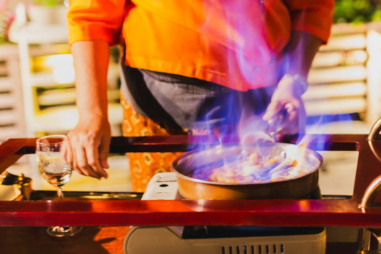 A person in an orange shirt flambés food in a pan on a portable stove, with blue flames rising and a glass of white wine nearby.