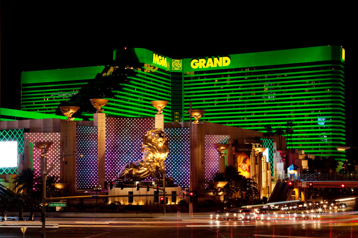 The MGM Grand Hotel in Las Vegas is brightly illuminated at night with green lights, featuring its iconic gold lion statue and light trails from passing traffic in the foreground.