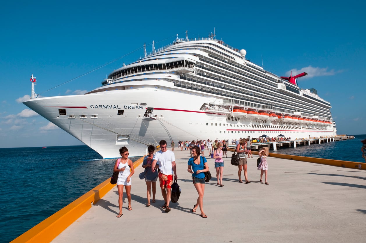 People walk on a sunny dock toward a large white cruise ship named Carnival Dream, which is docked at a pier over blue water under a clear sky.