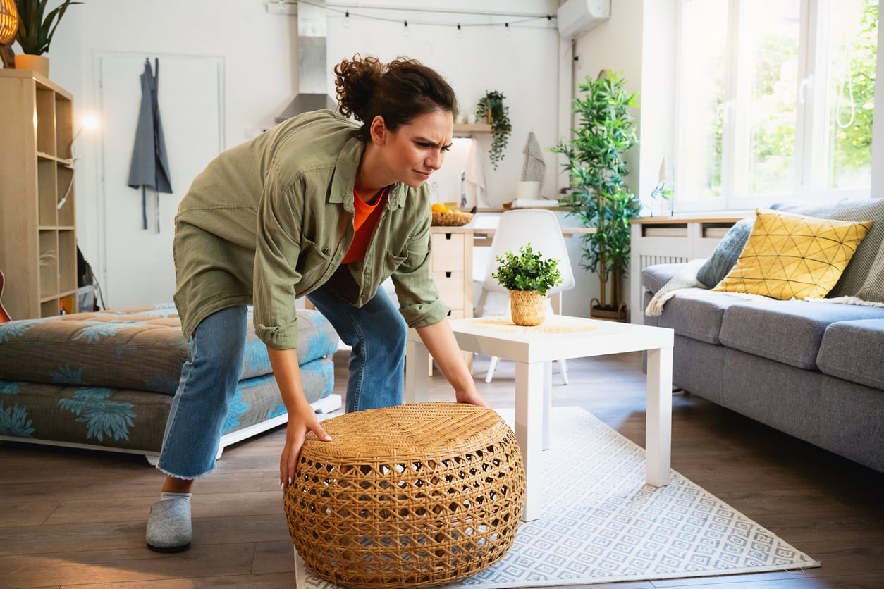 A person in casual clothes lifts one of the household items in a bright living room with a sofa, coffee table, and potted plants.