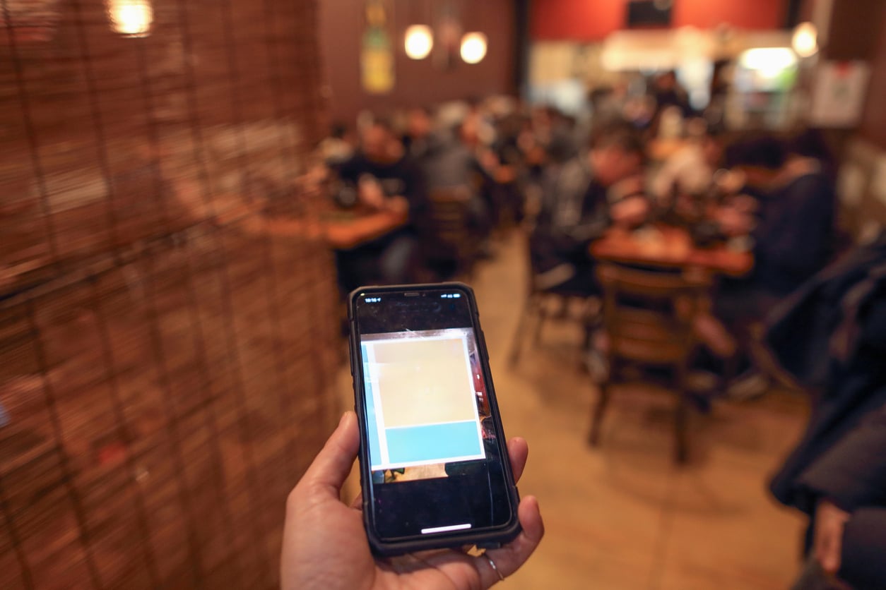 A hand holds a smartphone showing a blurred screen in focus, while the background reveals a busy, warmly lit restaurant with people seated at tables.