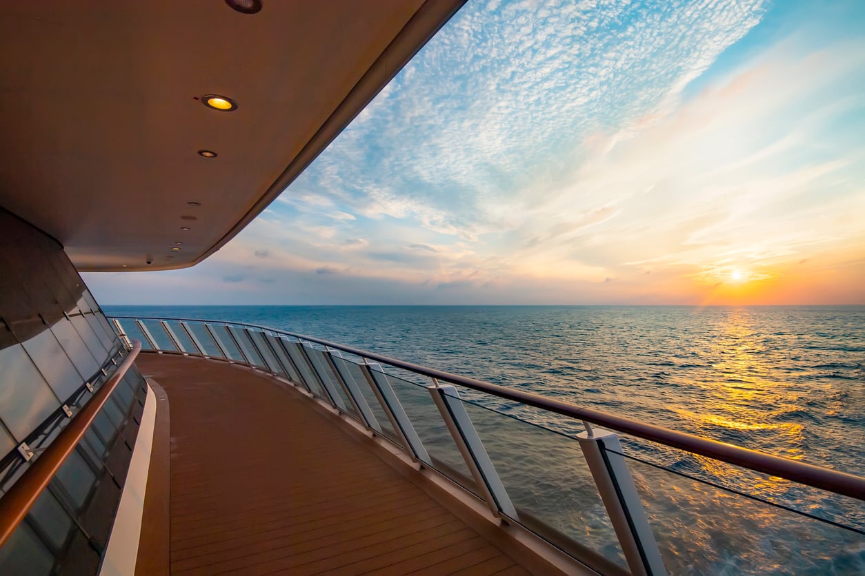 A view from a ship’s deck shows the ocean and a colorful sunset, with clouds and the sun near the horizon. The ship’s railing and lights are visible on the left.