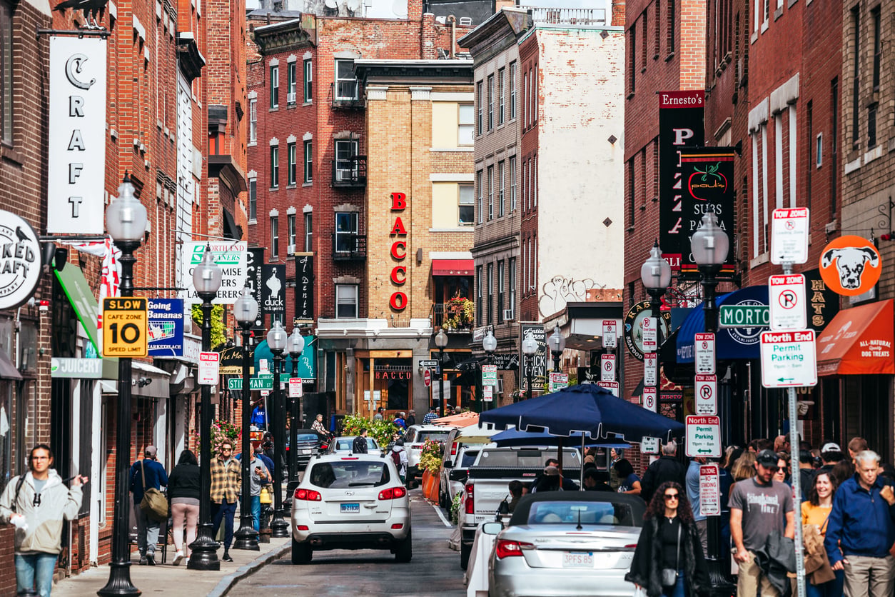 Busy city street lined with brick buildings, shops, restaurants, and signs; people walk on sidewalks while cars drive and park along the road under a bright, clear sky.