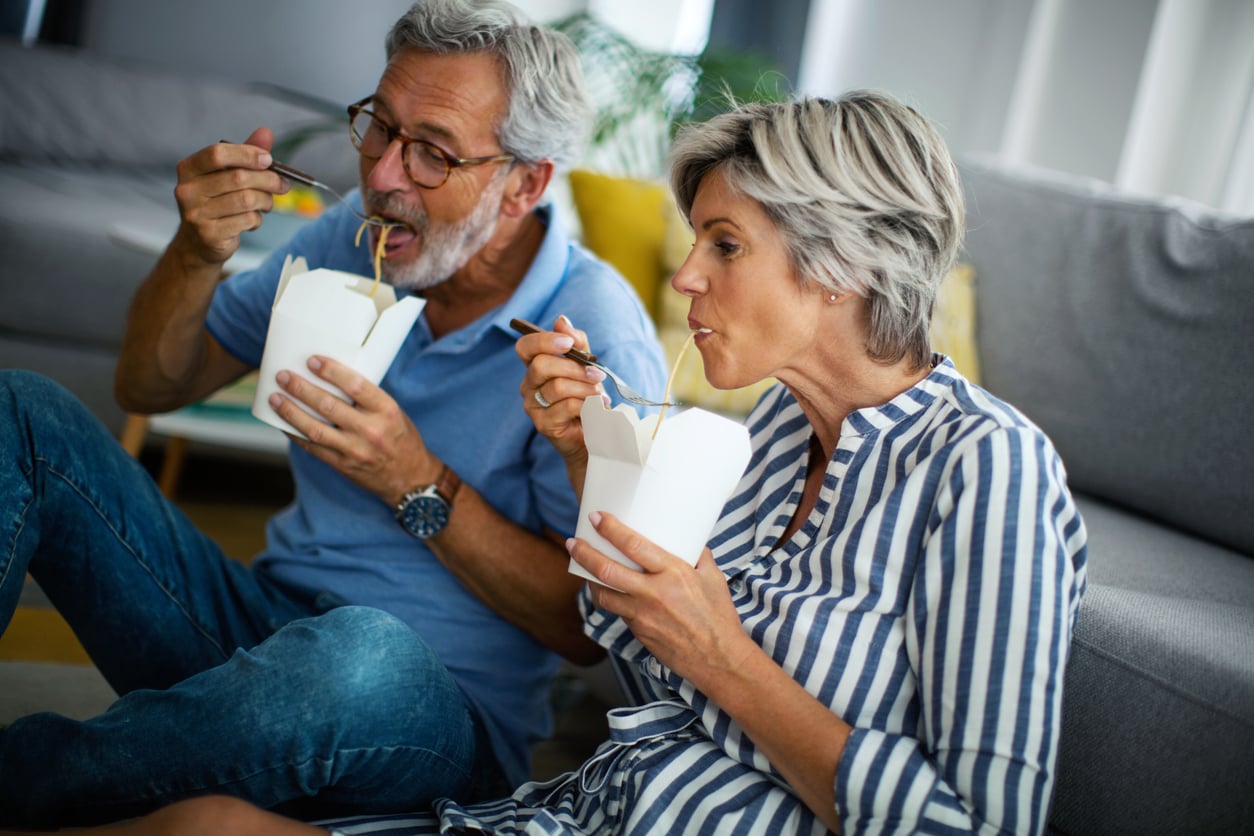 An older man and woman sit on the floor at home, enjoying noodles from white takeout boxes. Both are dressed casually and appear relaxed and happy as they eat together.