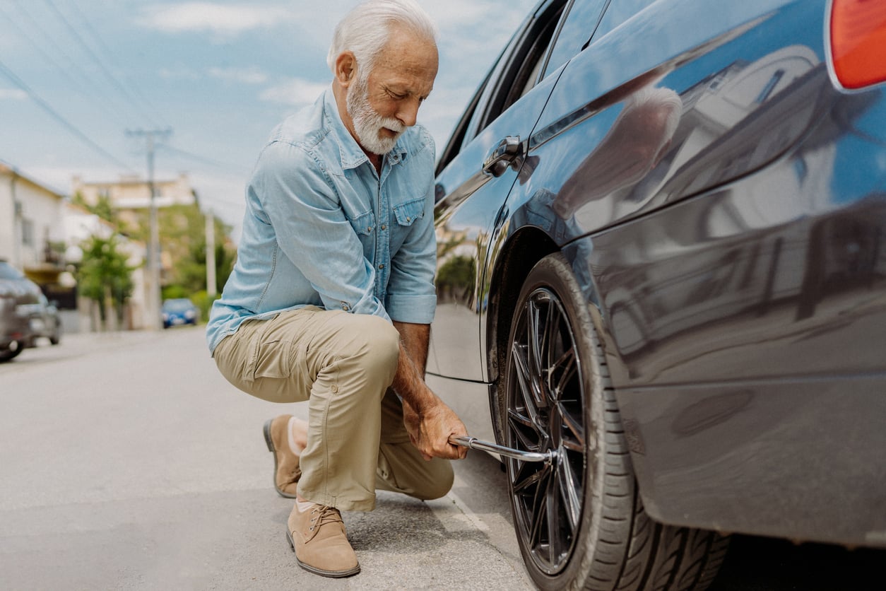 An older man with a gray beard is kneeling on the street beside a black car, using a tire iron to change the front tire.