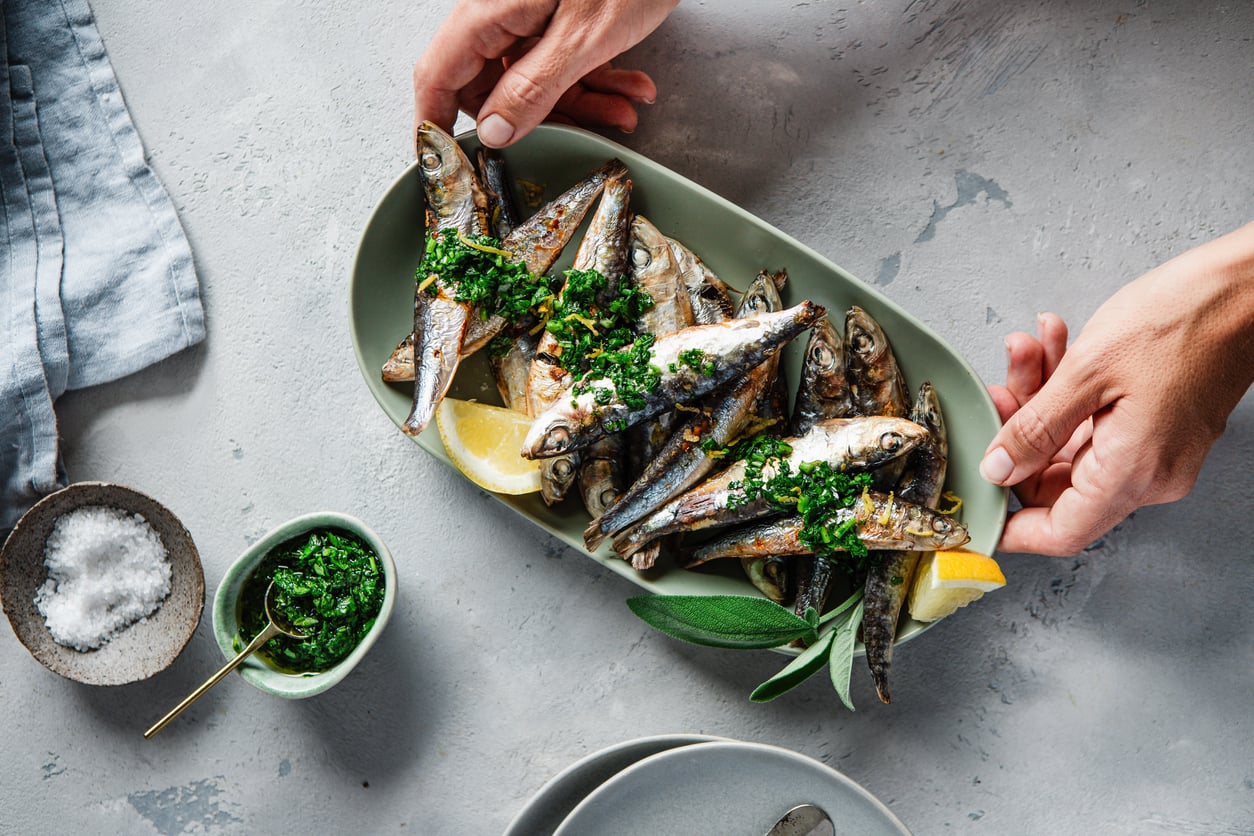 A person places a platter of grilled sardines garnished with chopped herbs and lemon wedges on a table. A bowl of salt, a small bowl of green sauce, and a napkin are nearby.