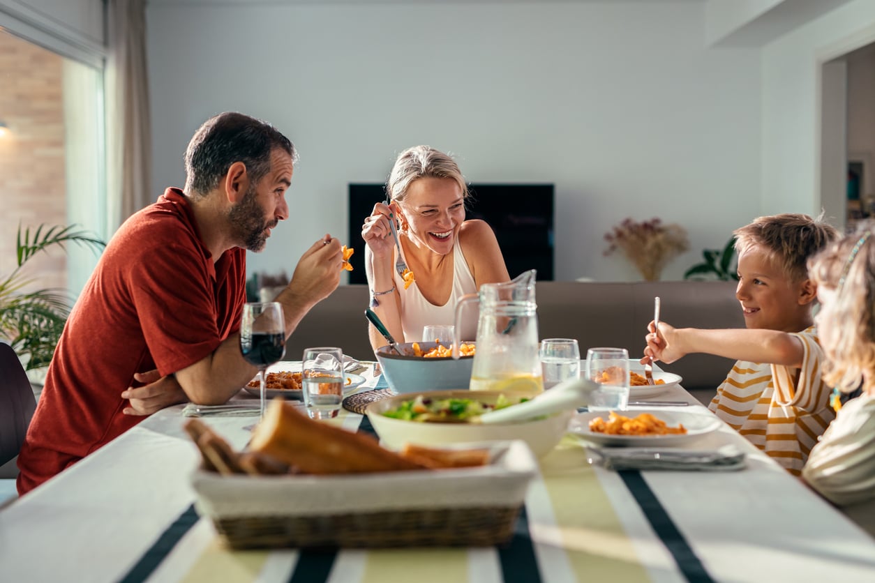 A family of four sits around a dining table, smiling and enjoying a meal together in a bright, modern dining room. Food and drinks are arranged on the table.