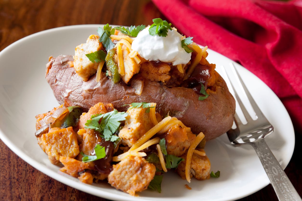 A baked sweet potato topped with seasoned tofu cubes, shredded cheese, cilantro, and a dollop of sour cream, served on a white plate with a fork and a red napkin.