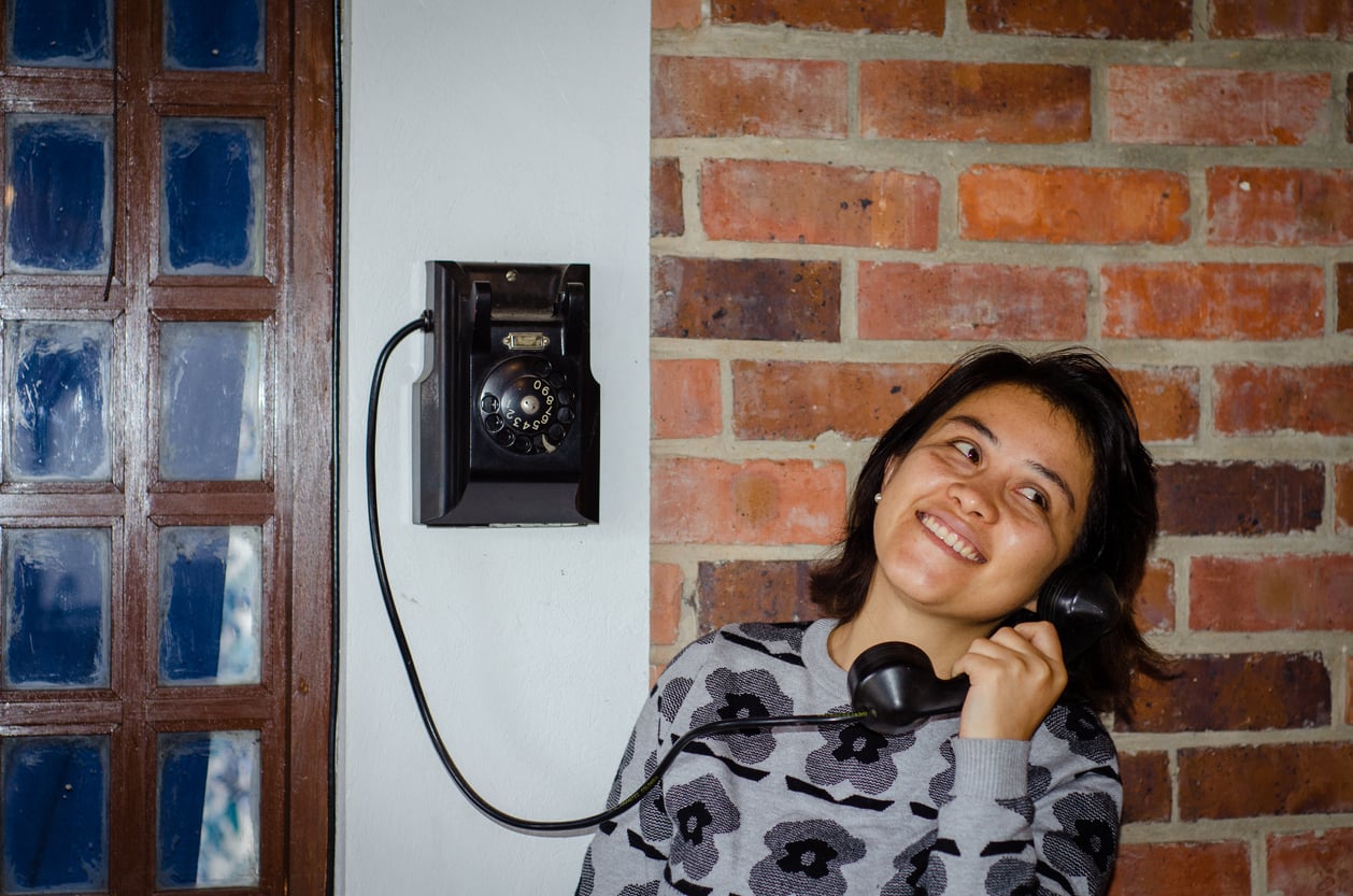 A person smiles while talking on a black rotary wall phone, standing against a brick wall and white section next to a wooden door with glass panes.