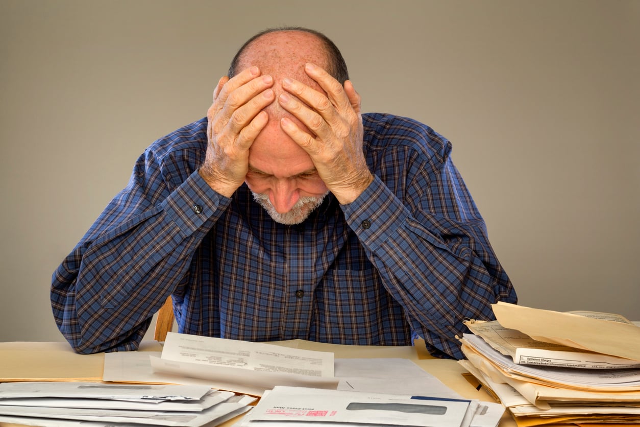 An older man in a plaid shirt sits at a table, holding his head in his hands, surrounded by paperwork and mail, appearing stressed or overwhelmed—perhaps anxious about his retirement money habits.