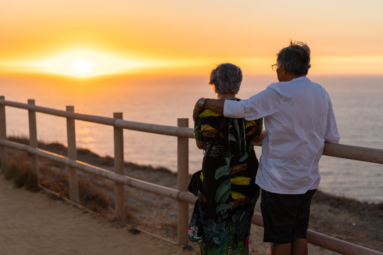 Two older adults stand arm in arm by a wooden fence, looking at the ocean and sunset. The sky glows orange, and they appear peaceful and content—cherishing one of their favorite vacation experiences together.