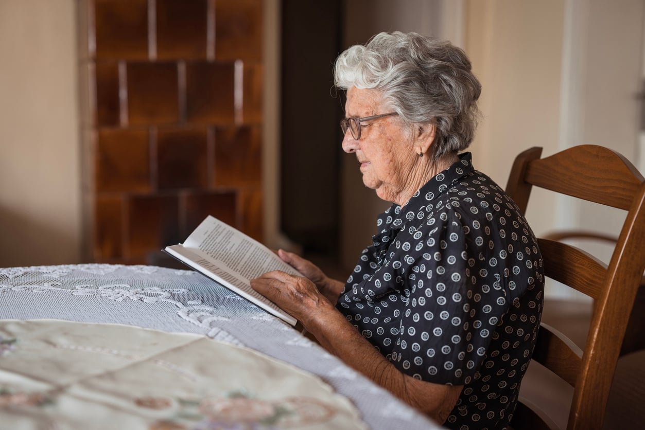An elderly woman with gray hair and glasses sits at a table, reading a book. She wears a black dress with white polka dots. Daylight comes from the side, softly lighting the scene.