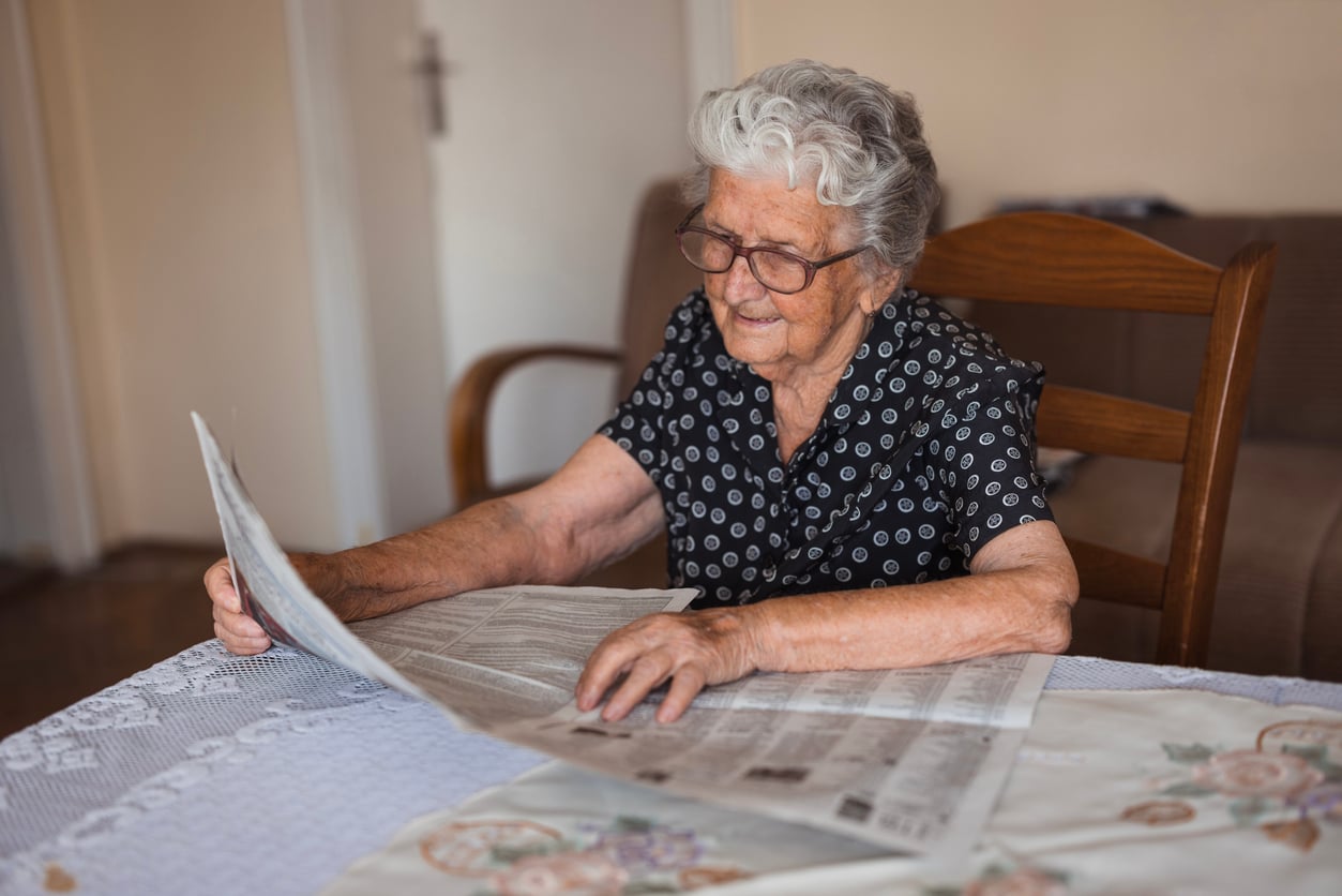 Elderly woman with glasses and gray hair sits at a table covered with a lace tablecloth, reading a newspaper. She wears a patterned black blouse and smiles gently in a cozy room.