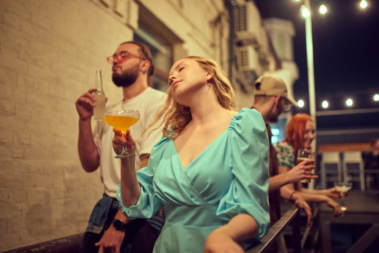 A woman in a blue dress holds a drink and dances at an outdoor gathering at night, with three other people nearby under string lights.
