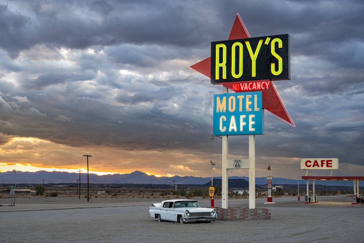 A classic white car is parked near a large retro sign reading “Roy’s Motel Café” under dramatic, cloudy skies at sunset in a remote desert setting. The empty parking lot and café evoke a nostalgic, lonely atmosphere reminiscent of travel in the 60s and 70s.