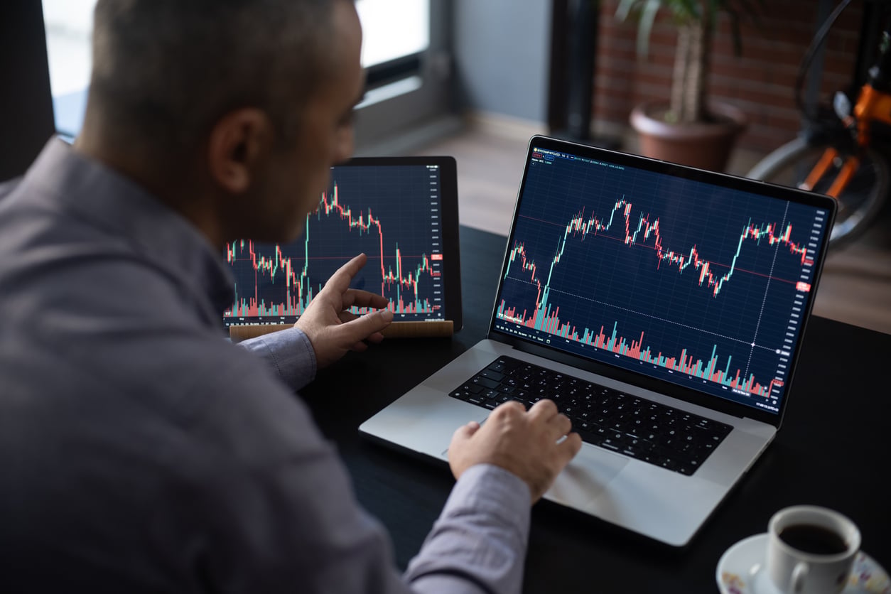 A person analyzes financial charts on a tablet and a laptop at a desk, both displaying colorful stock market graphs and candlestick patterns.