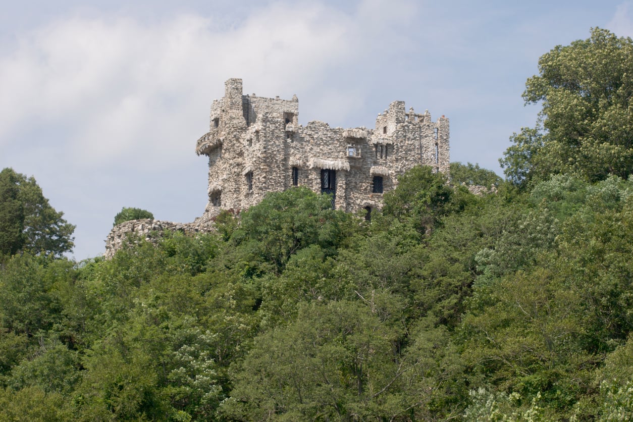A partially ruined stone castle stands atop a hill, surrounded by dense green trees, with a blue sky and some clouds in the background—a striking example of how castles gracefully blend history with nature.