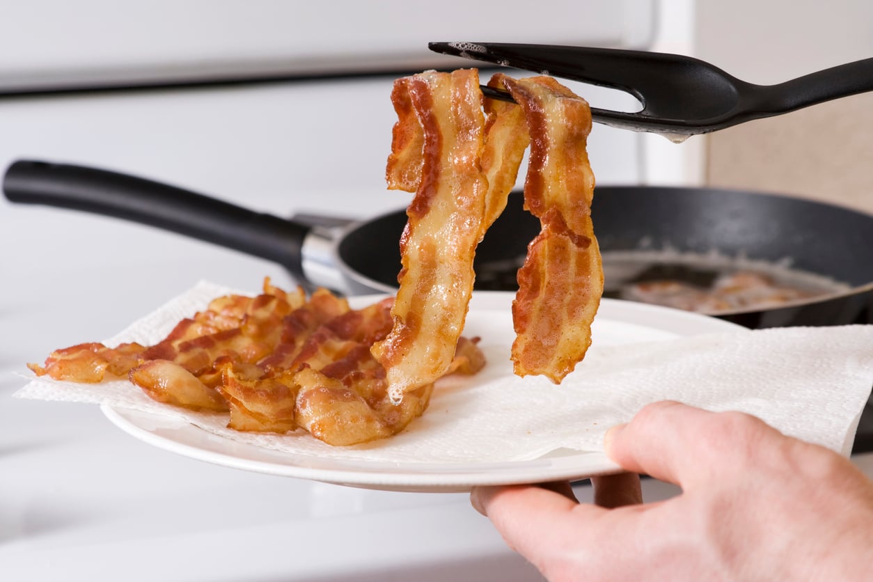 A hand uses tongs to place crispy cooked bacon strips onto a white plate lined with paper towels, with a frying pan and stove in the background.