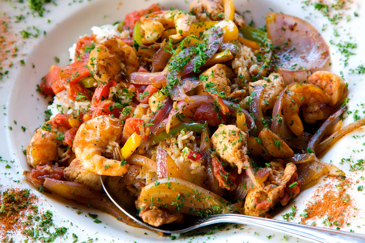 A close-up of a plate filled with rice, shrimp, chicken, sautéed onions, bell peppers, and herbs, with a spoon on the side and seasoning sprinkled around the edge of the plate.