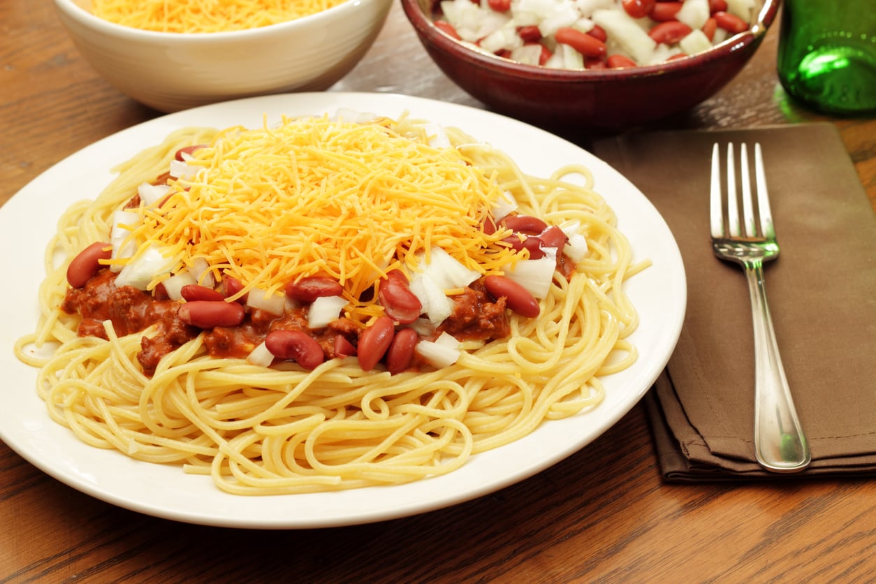 A plate of spaghetti topped with chili, kidney beans, diced onions, and shredded cheddar cheese, with a fork and napkin beside it. Bowls of cheese and chopped onions are in the background.