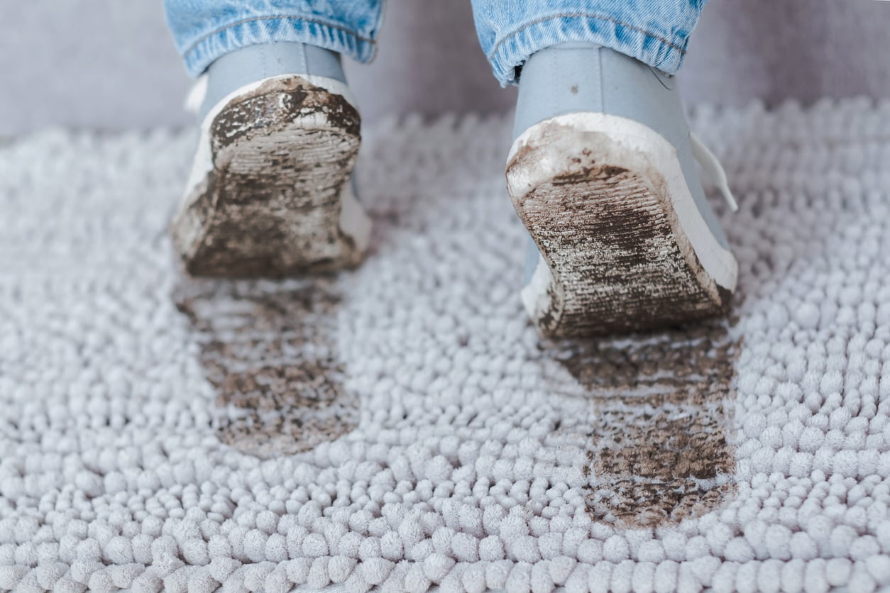A person wearing muddy shoes stands on a light gray carpet, leaving wet and dirty footprints behind them. The photo is taken from behind, focusing on the shoes and the stains.
