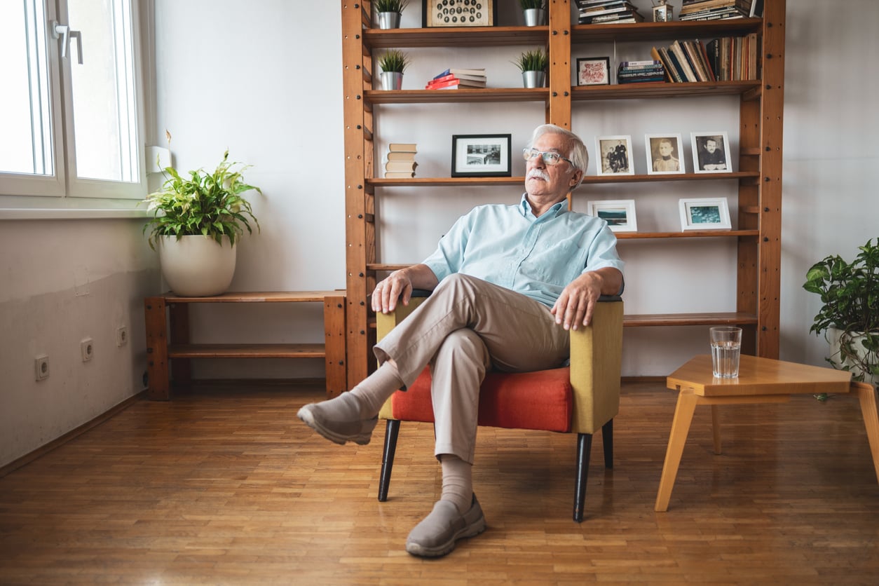 An older man with gray hair and glasses sits in a colorful armchair in a cozy room with wooden floors, bookshelves, potted plants, and a small table with a glass of water.