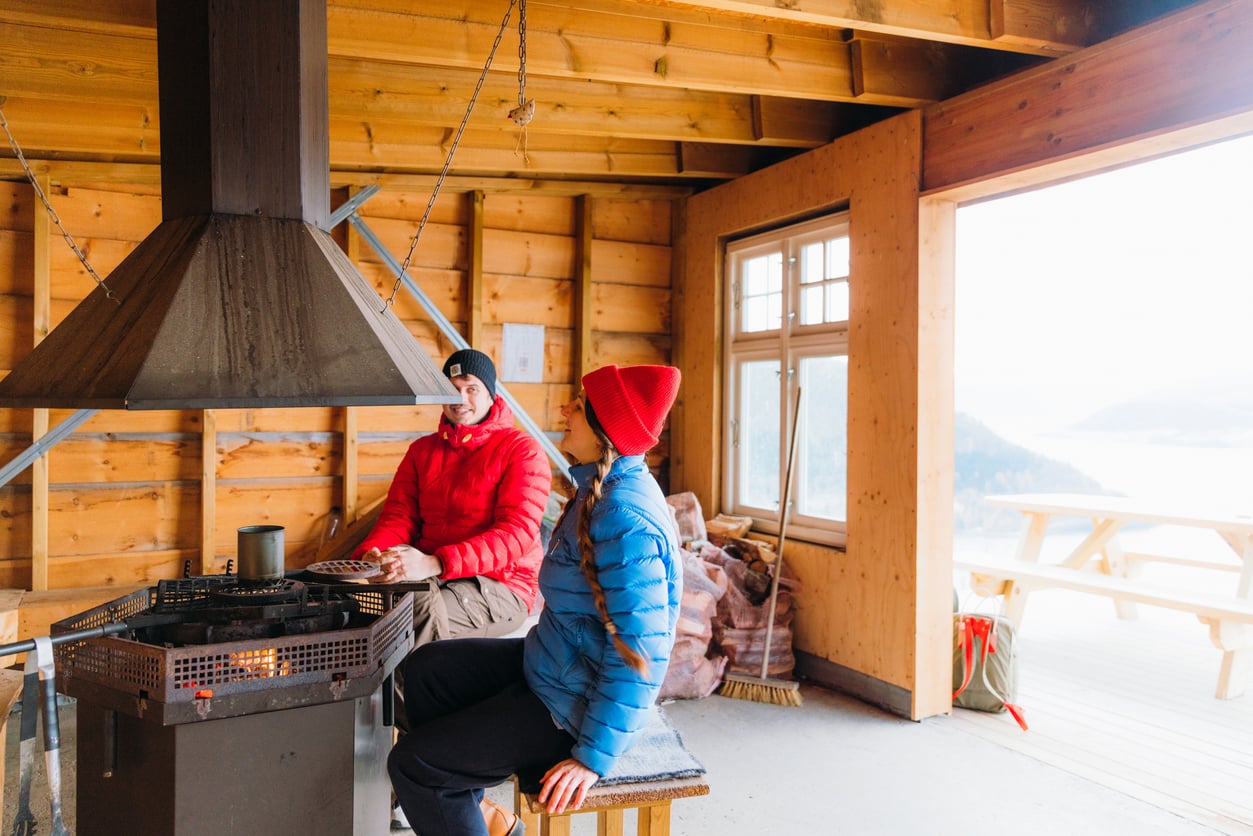 Two people in winter jackets sit by an indoor fire pit in a wooden cabin with large windows, enjoying the warmth. A snowy outdoor scene with picnic tables is visible through the open doorway.