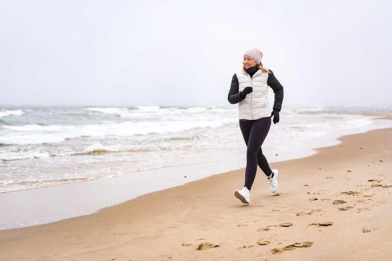 A woman in a white jacket and gray hat jogs along a sandy beach near the ocean, leaving footprints behind her. The sky is overcast and the waves are gently crashing onto the shore.