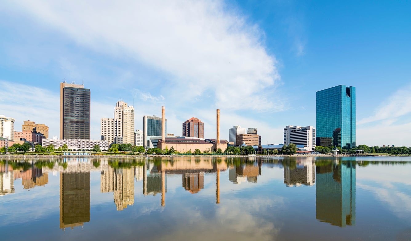 A city skyline with tall buildings reflected in a calm river under a bright blue sky with wispy clouds.