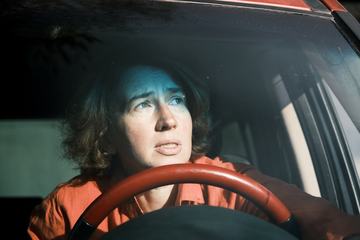 A worried person with short brown hair sits behind the wheel of a car, looking through the windshield while sunlight casts shadows on their face.