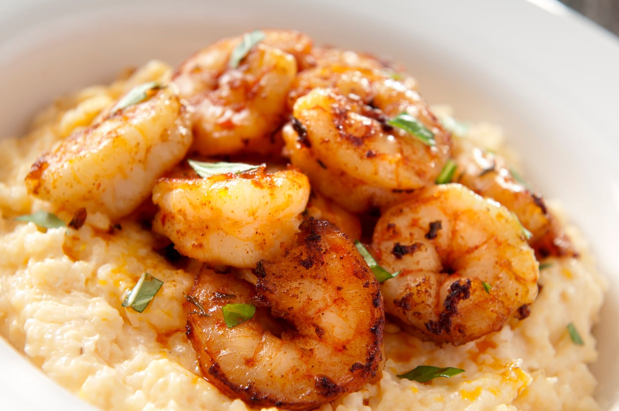A close-up of creamy grits, a classic Southern foods favorite, topped with seasoned, grilled shrimp and garnished with chopped green herbs, served in a white bowl.