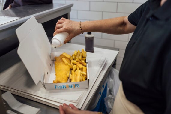 A person sprinkles salt on a box of fish and chips&mdash;one of the classic foods to order&mdash;in a takeaway container, standing by a counter in a tiled kitchen.