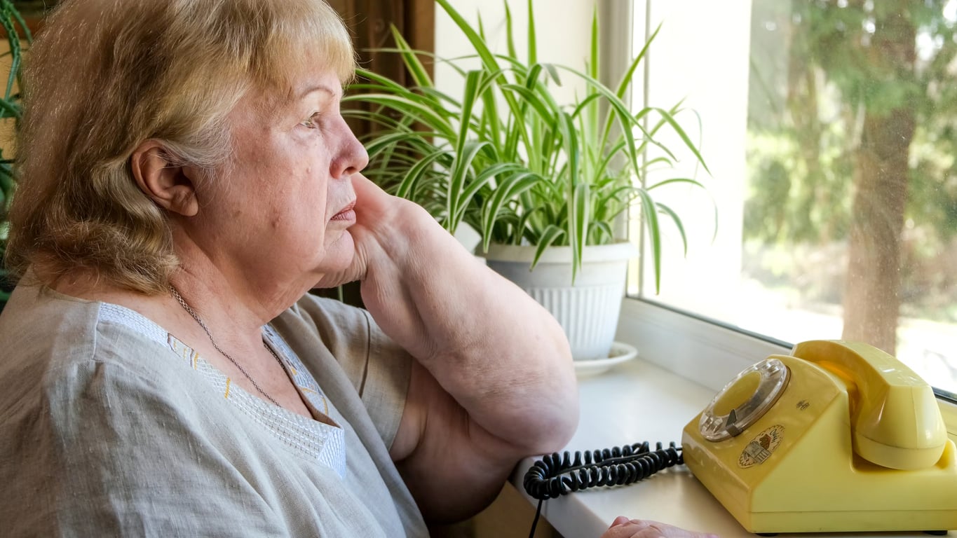 An older woman sits by a window, resting her hand on her neck, gazing outside. A yellow rotary phone and a potted plant are on the windowsill beside her.