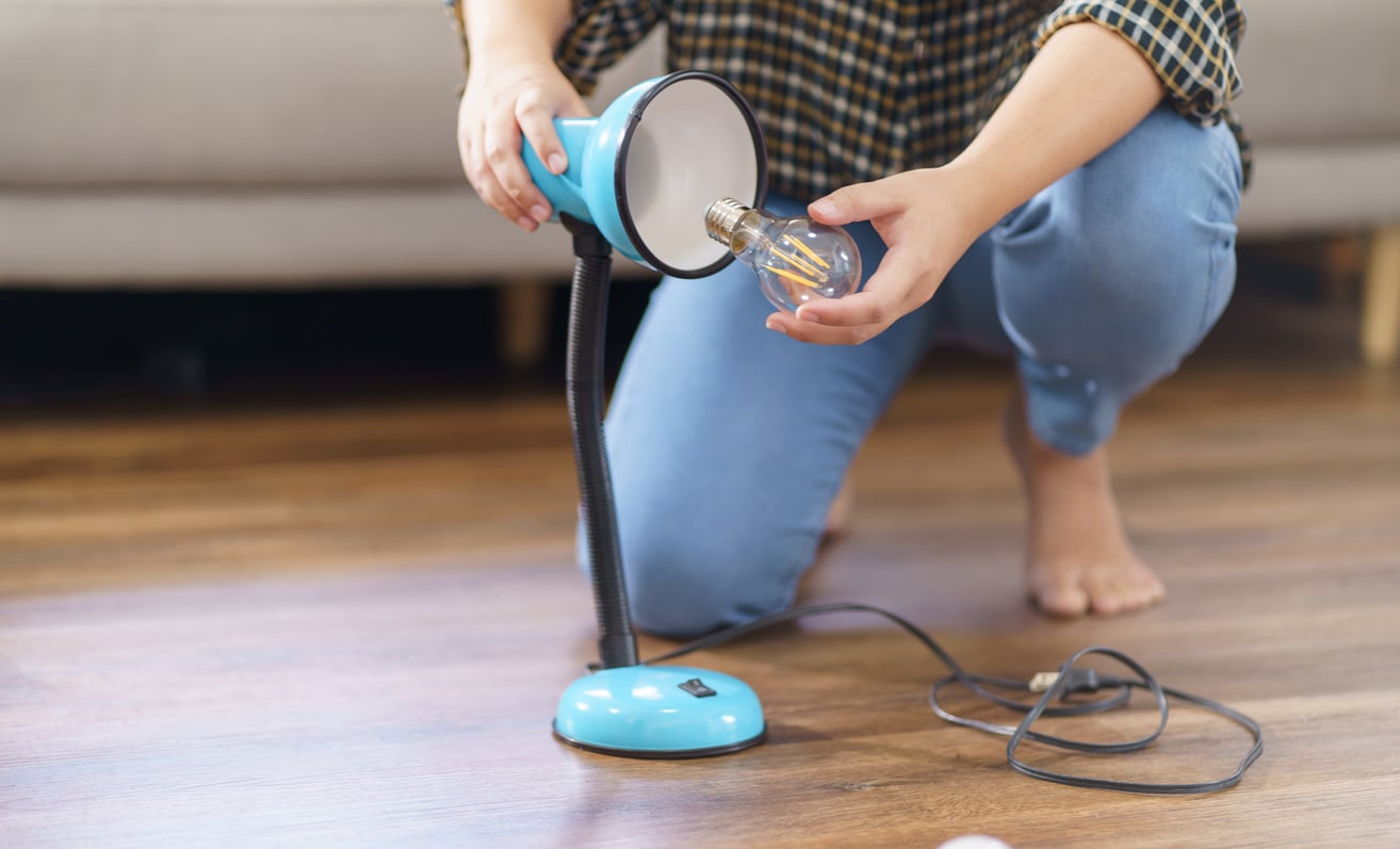 A person kneels on a wooden floor, holding a blue desk lamp and inserting a lightbulb into its socket. The lamp’s cord is lying on the floor nearby.