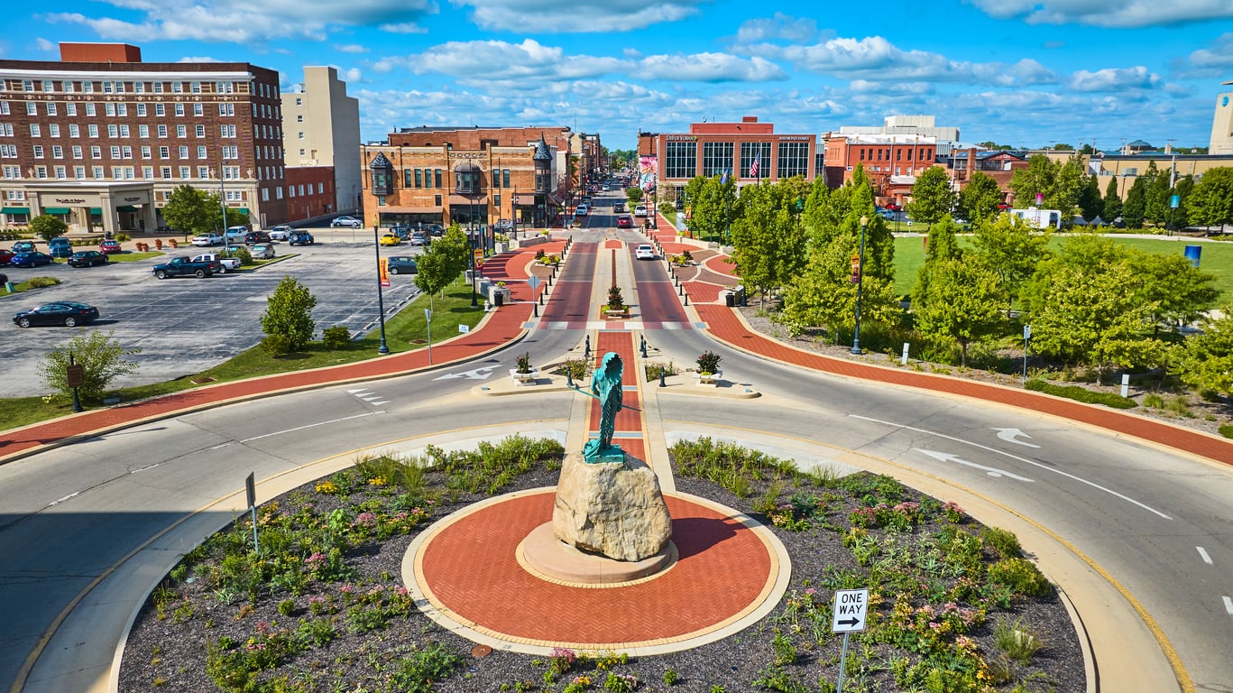 A roundabout with a statue on a rock in the center, surrounded by greenery, leads to a downtown area with red brick buildings, parked cars, and a bright blue sky with scattered clouds.