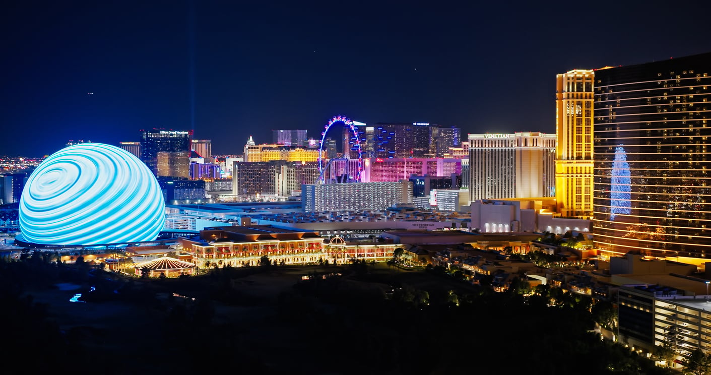 Las Vegas skyline at night featuring the illuminated MSG Sphere with blue swirling lights, colorful hotels, the High Roller Ferris wheel, and bright city lights against a dark sky.