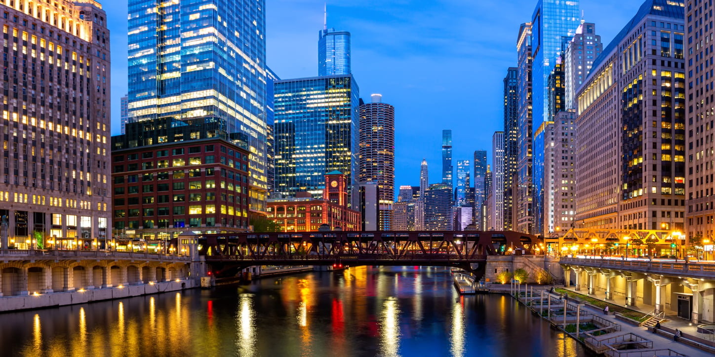 City skyline at dusk with illuminated skyscrapers reflecting on a river, a bridge crossing the water, and lights glowing along the buildings and waterfront—an inviting scene that captures the perfect atmosphere for a memorable vacation.