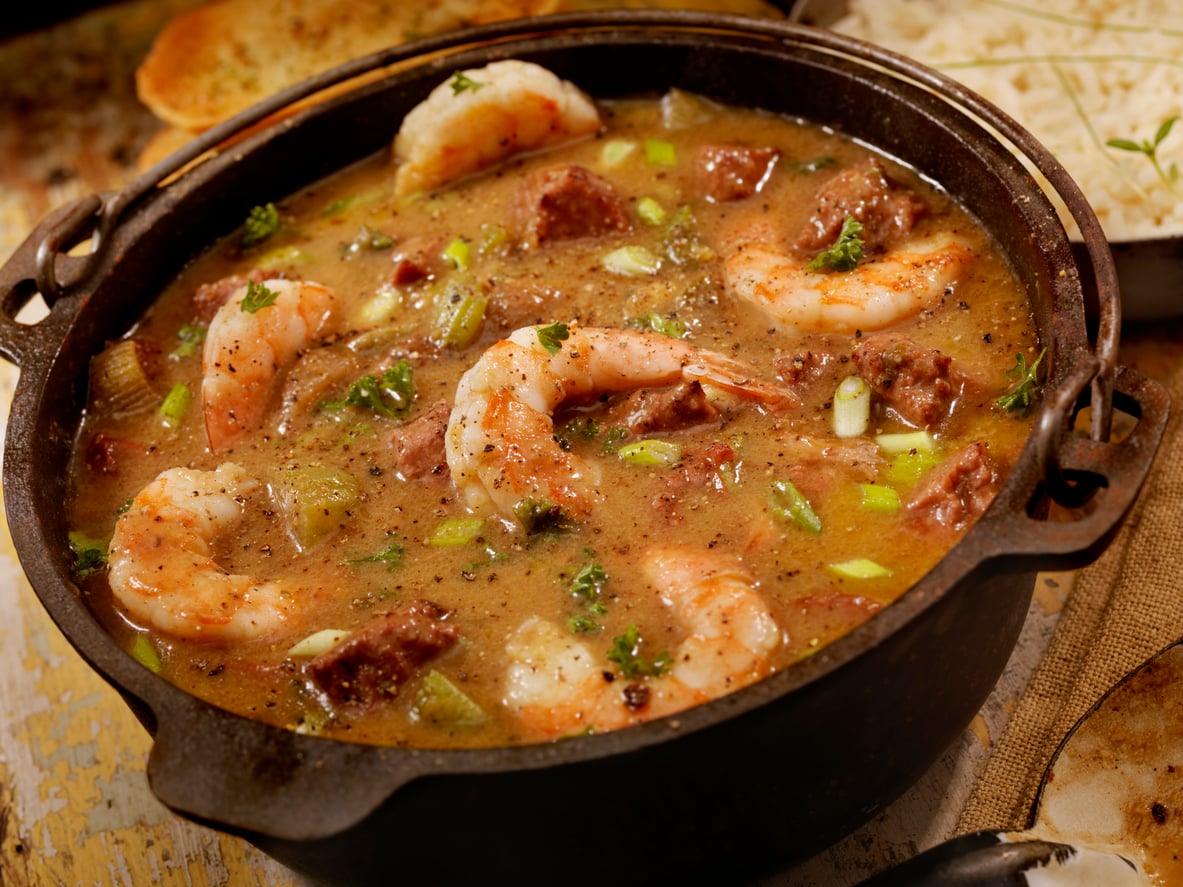 A pot of gumbo filled with shrimp, sausage, green onions, and herbs in a rich, brown broth sits on a rustic table.