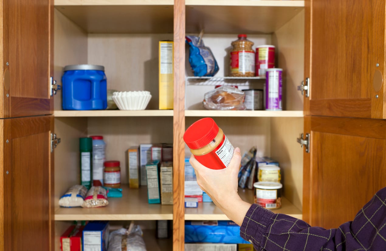 A person holds a peanut butter jar while looking into a kitchen pantry filled with various food items, including jars, cans, and boxes, on wooden shelves.