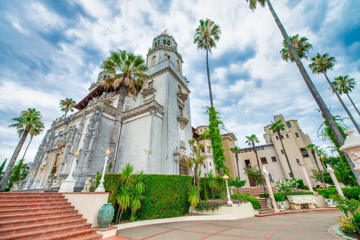 A grand historic mansion reminiscent of elegant castles, with ornate towers, surrounded by palm trees and lush greenery under a cloudy blue sky. Wide brick steps lead to the entrance.
