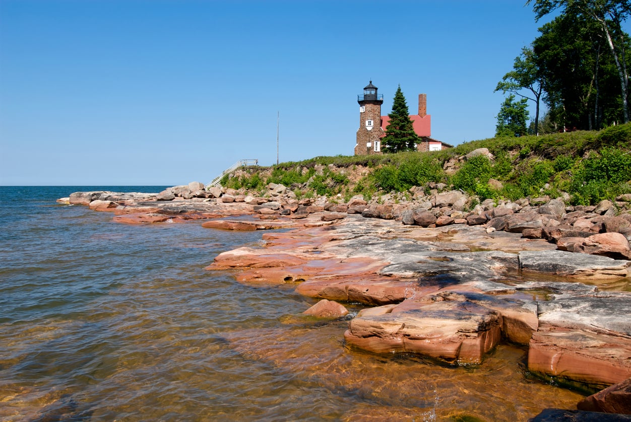A lighthouse sits on a rocky, tree-lined shoreline overlooking a large body of water under a clear blue sky.