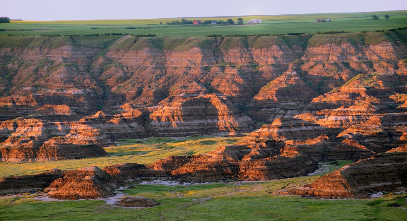 Sunlight hits layered, eroded rock formations in a canyon, with grassy fields and a few houses on top of the cliffs in the background, making it one of the region’s most stunning hiking destinations.