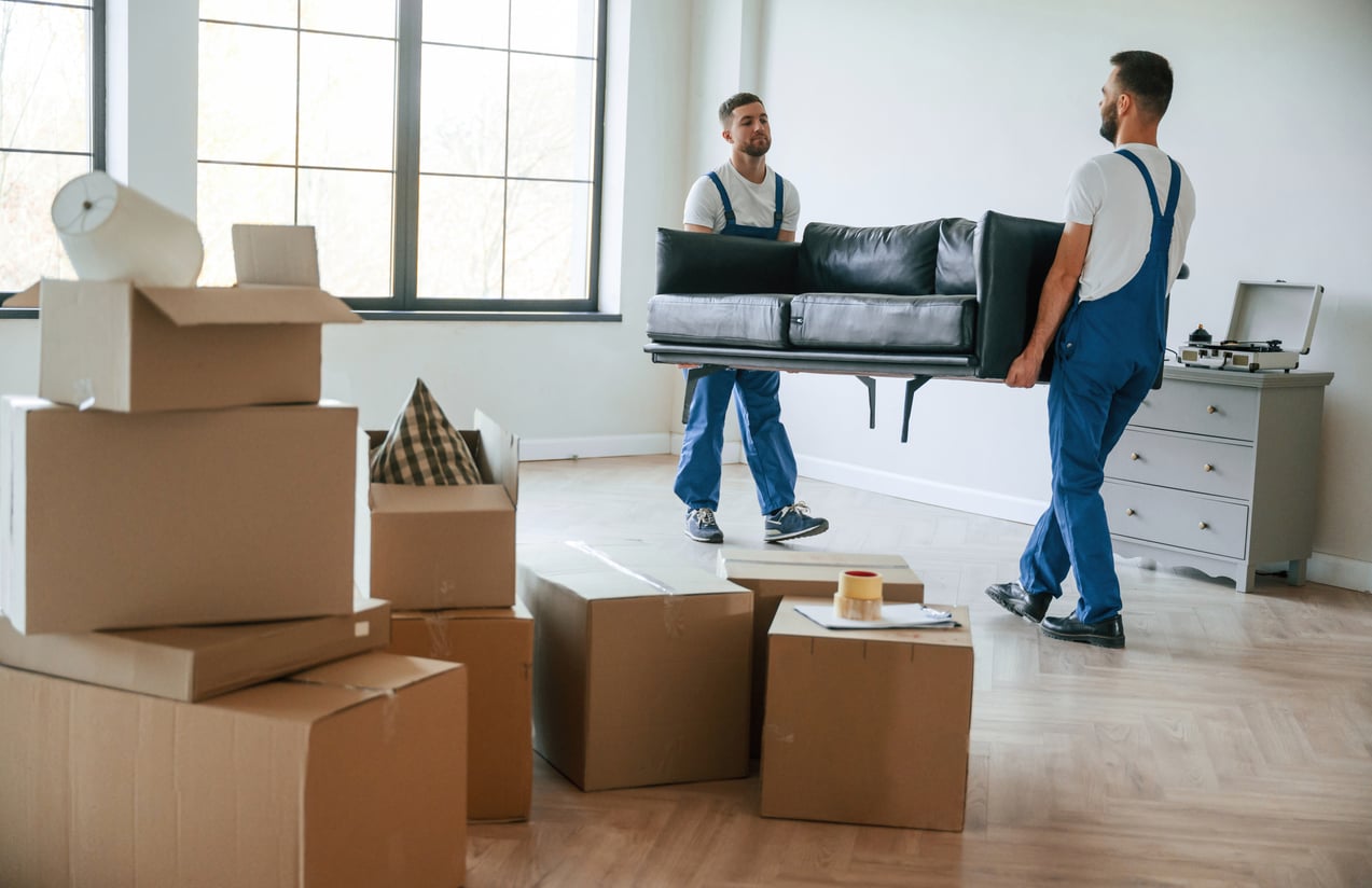 Two men in blue overalls are carrying a black sofa in a bright room with large windows. Cardboard boxes and packed items are scattered on the floor, suggesting a move or relocation.
