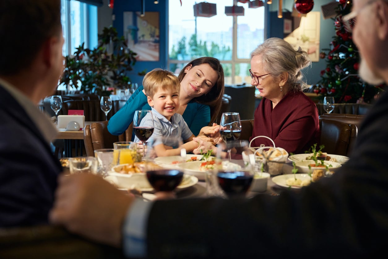 A family sits around a festive table, smiling and enjoying a meal together. A young boy laughs while a woman and an elderly lady sit beside him. Holiday decorations and a tree are visible in the background.